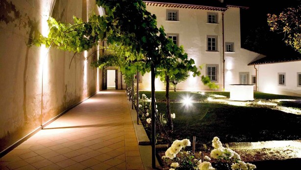 En korridor upplyst under natten, med ett klinkergolv, en vit byggnad med många fönster och en trädgård med vita blommor och träd.