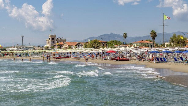 A beach with umbrellas and lounge chairs, a boat, and a building with a flag.