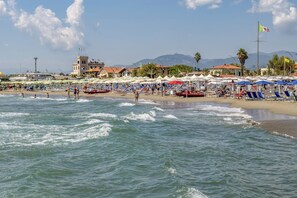 A beach with umbrellas and lounge chairs, a boat, and a building with a flag. - Holiday apartment with air conditioning and outdoor pool (Marina di Massa)