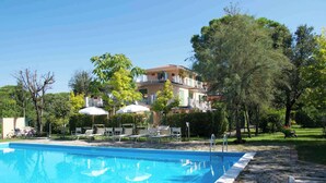 A swimming pool with lounge chairs and umbrellas, surrounded by trees and a building with balconies.