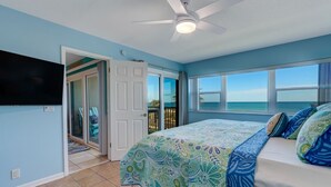 A bedroom with a bed, a ceiling fan, a TV mounted on the wall, and a view of the ocean through large windows. - Beachfront Beauty: Condo with Pier Access! (Fernandina Beach)