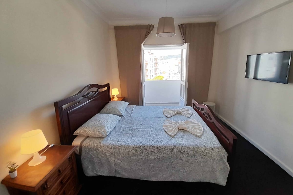 Ein Schlafzimmer mit Bett, Nachttischlampen, einem Schminktisch, einem Fernseher und Blick ins Freie.