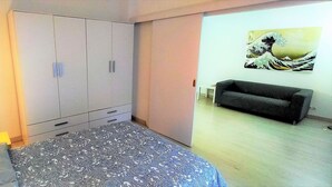 A bedroom with a bed, wardrobe, bedside lamp, and a sofa in the adjacent room. - Beautiful furnished studio of 67 m2 (Barcelona)
