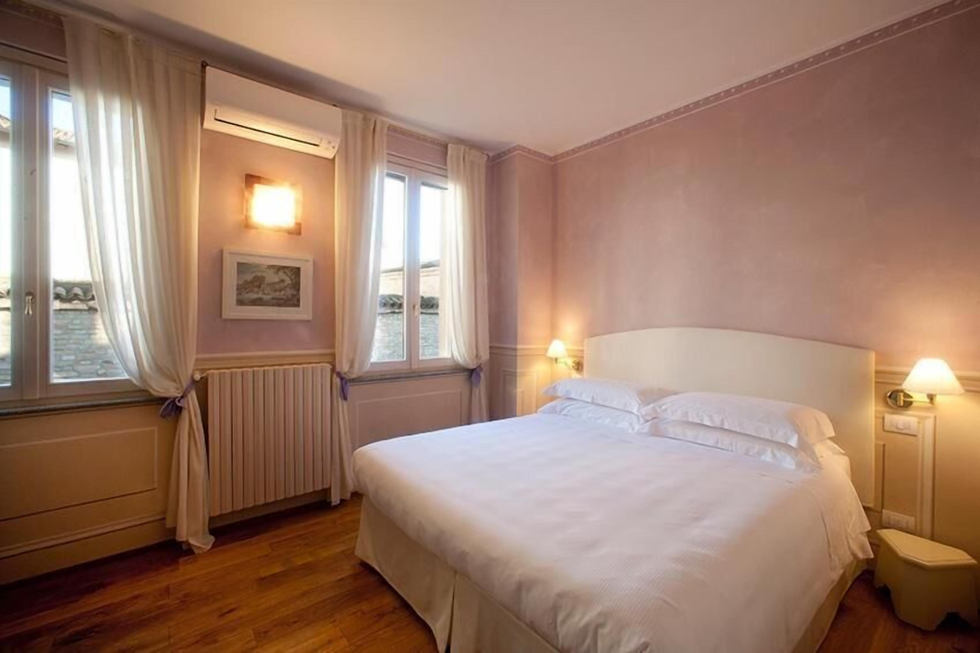 Habitación de hotel con una cama grande, dos lámparas de noche, un radiador y una ventana con cortinas.