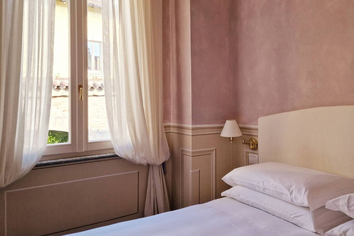 A hotel room with a bed, white curtains, a window, and a wall-mounted lamp.