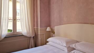 A hotel room with a bed, white curtains, a window, and a wall-mounted lamp.