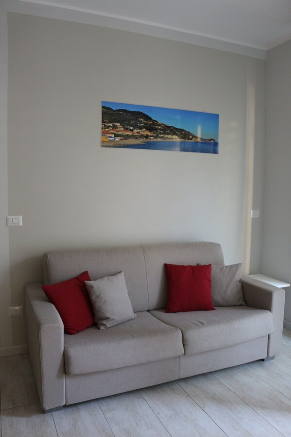 A neutral-toned sofa with two red cushions, a small white side table, and a framed landscape painting on the wall.