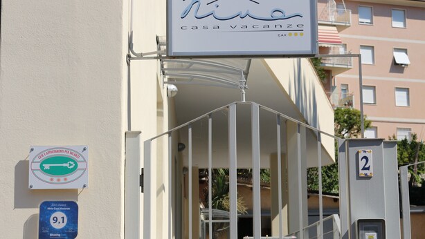 Une entrée fermée par un portail mène à un immeuble. Une plaque indique « Nina Casa Vacanze », et un numéro « 2 » est affiché sur un poteau en métal.