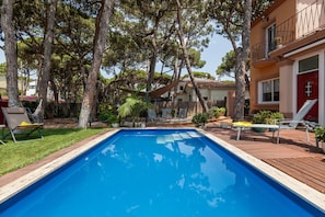 A rectangular swimming pool with a wooden deck surrounding it, trees, and a building with a red door.