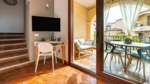 Ein Zimmer mit Schreibtisch, Stuhl, Fernseher und einem Balkon mit Sitzgelegenheit im Freien.