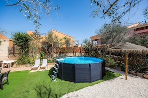 Un jardin avec une piscine, une échelle, une structure recouverte de chaume et une petite table avec des chaises.