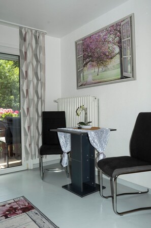 A room with a dining table, two chairs, a radiator, and a window with a view of trees and flowers. - Vacation apartment Sonnenblick (Oberharmersbach)