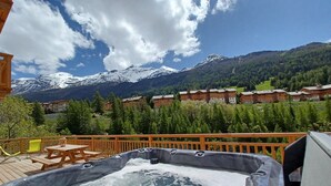 A wooden deck with a hot tub, a table, and chairs, overlooking a mountain range with snow-capped peaks and a forested valley. - Apartment 'Chalet La Turra' with Mountain View, Wi-Fi and Air Conditioning (Lanslebourg Mont Cenis)