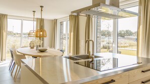 A modern kitchen with a large island, stainless steel appliances, and large windows offering a view of the outdoors. - Optoog XL (Callantsoog)