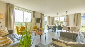 A modern living room with a sofa, a dining table, and chairs. There is a large window providing natural light and a view of the outdoors. - Optoog XL (Callantsoog)