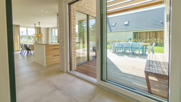 A modern kitchen with a central island, a dining area with a table and chairs, and a large sliding glass door leading to an outdoor patio with a wooden deck and a table set for dining. - Optoog XL (Callantsoog)