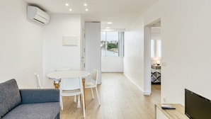 Una sala de estar moderna con un sofá gris, una mesa de comedor blanca con sillas, un televisor de pantalla plana y un aire acondicionado instalado en el techo.