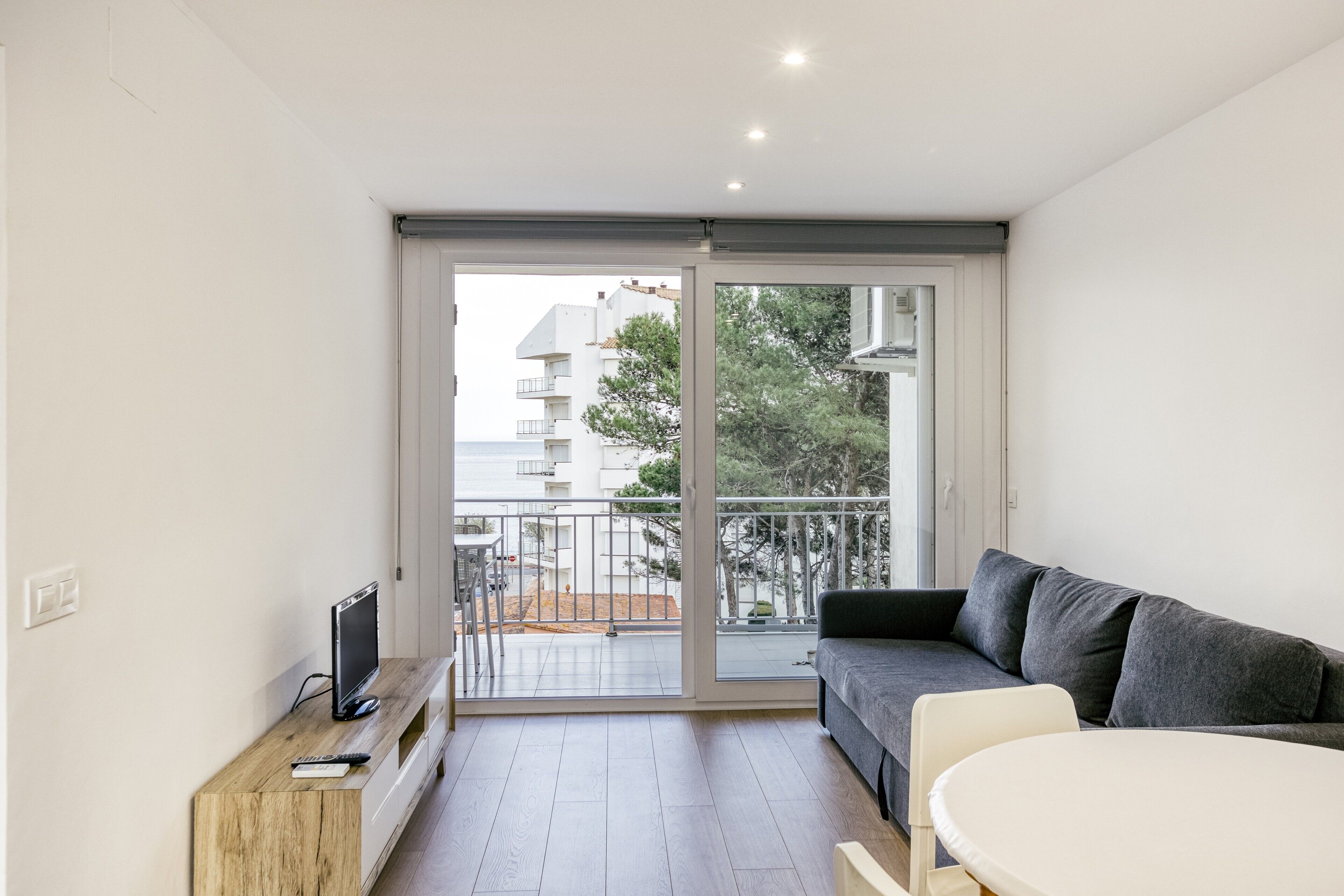 Una sala de estar moderna con un sofá, un televisor sobre un mueble de madera, una mesa de comedor y un ventanal corredero que da a un balcón.