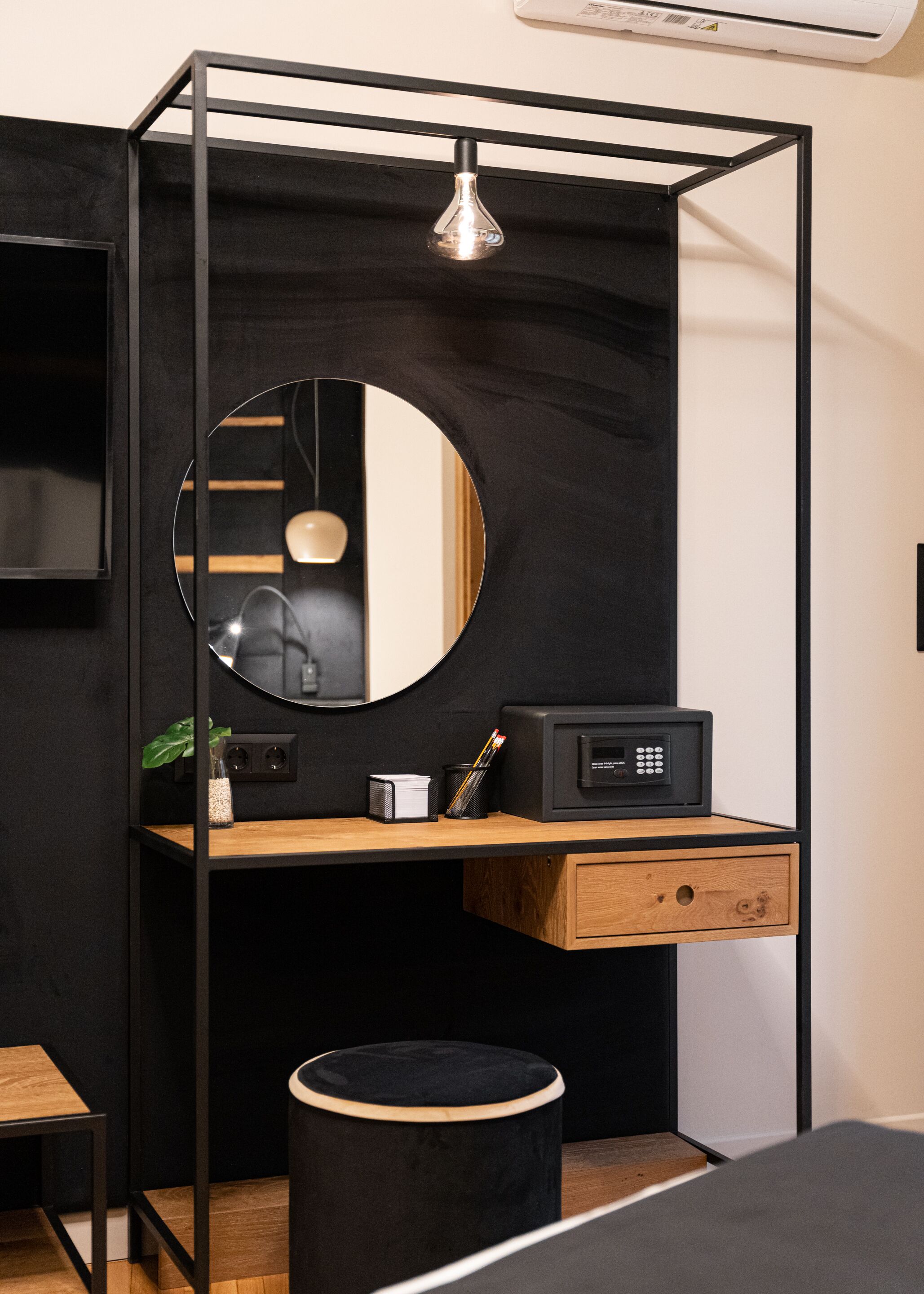 A compact, modern room with a black and wooden desk, a round mirror, a small black ottoman, and a black-framed bed.
