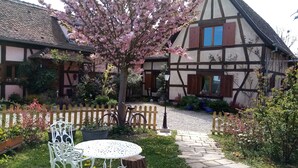 Una casa tradicional d'estuc i fusta amb un jardí que inclou una taula i cadires de metall blanc, una tanca de fusta i arbres en flor.
