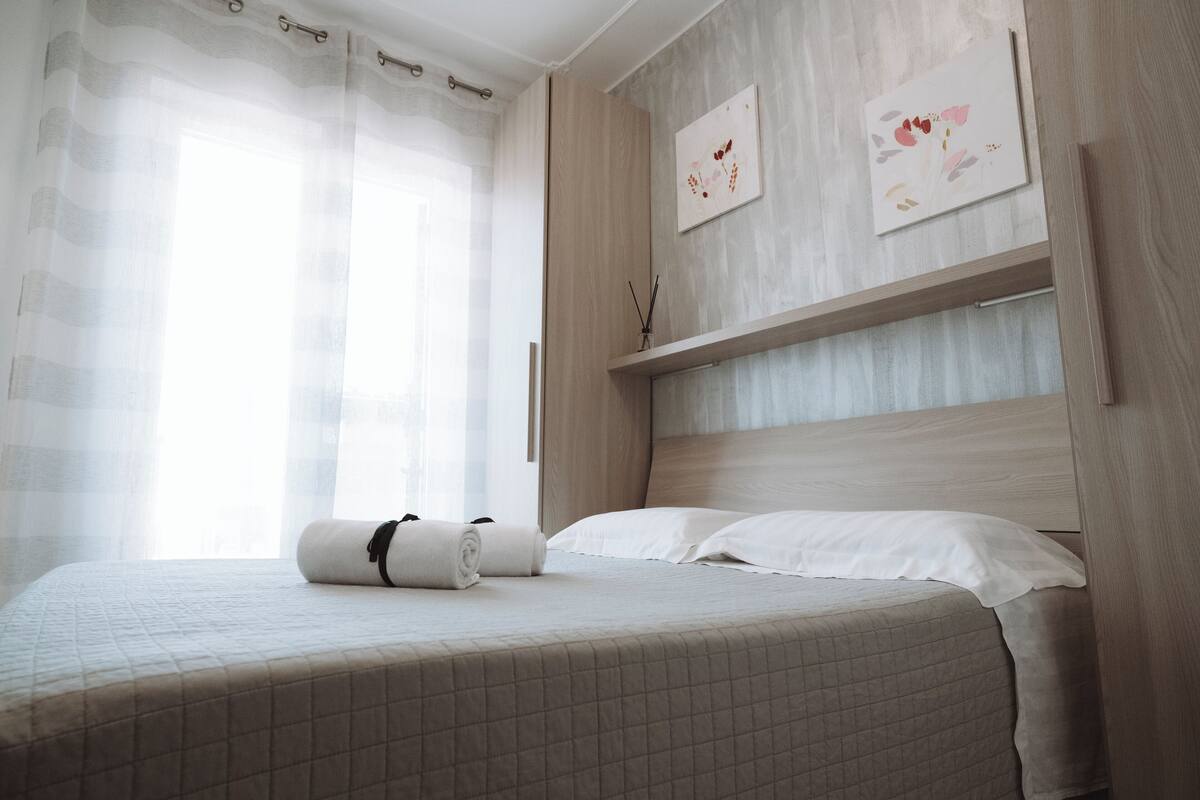 Cama bem arrumada com colcha acolchoada, toalha enrolada e cabeceira com prateleira acima.