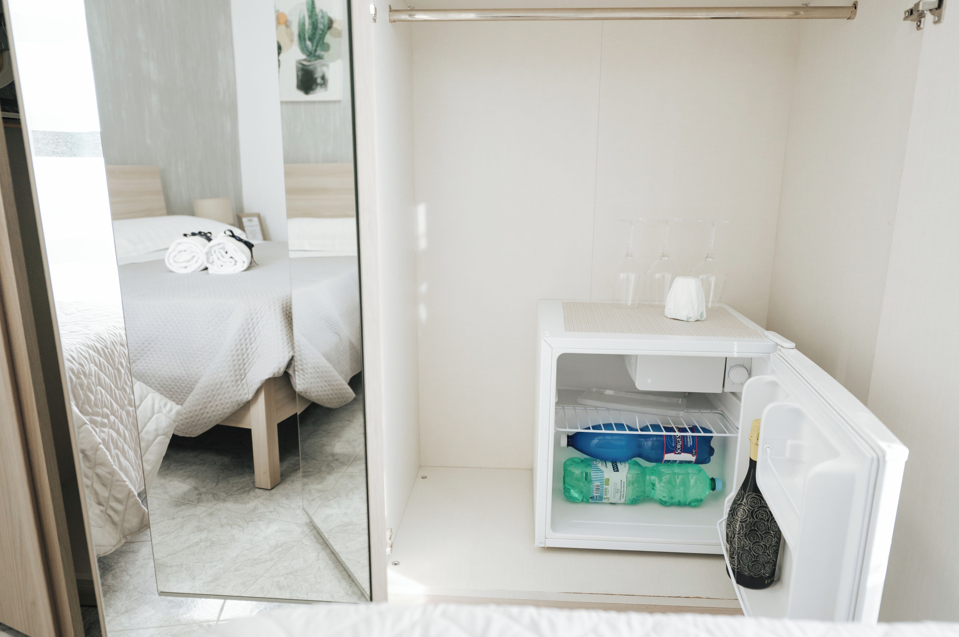 Una camera d'albergo con un letto, un bagno piccolo e un frigorifero con acqua in bottiglia e una bottiglia di champagne.