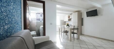 Una sala de estar moderna con un sofá gris, un área de comedor con mesa y sillas, y una cocina al fondo.
