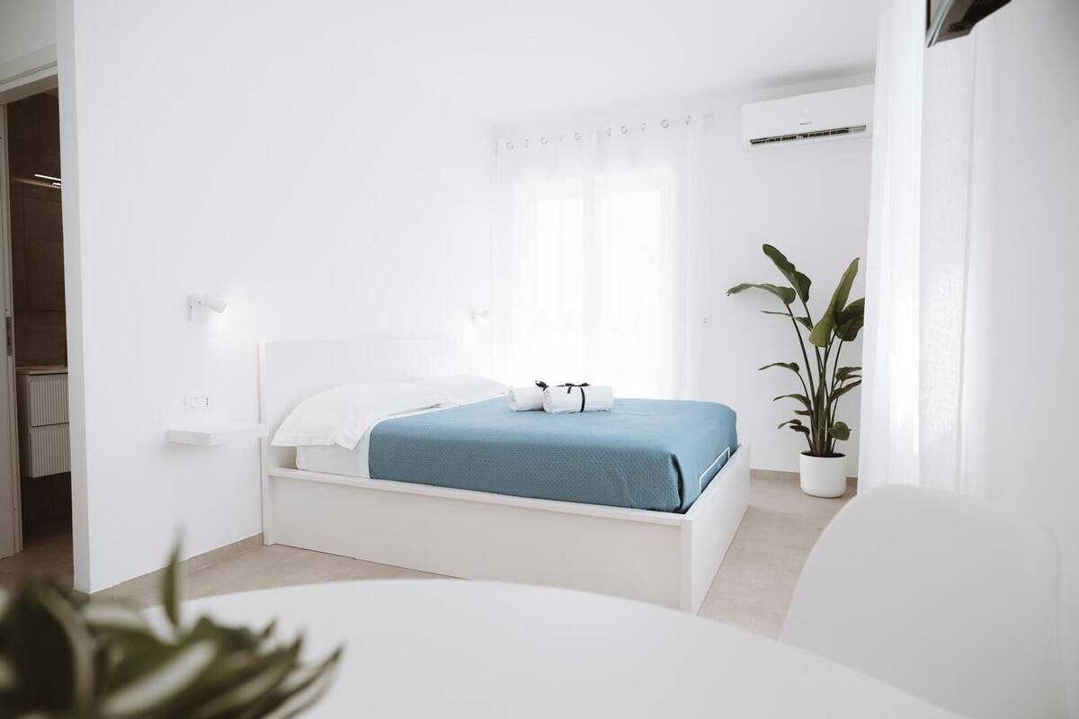 Quarto minimalista com uma cama, um vaso de planta e um guarda-roupa branco.