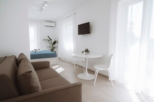 A modern hotel room with a bed, a sofa, a dining table with chairs, a TV mounted on the wall, and large windows with sheer curtains.