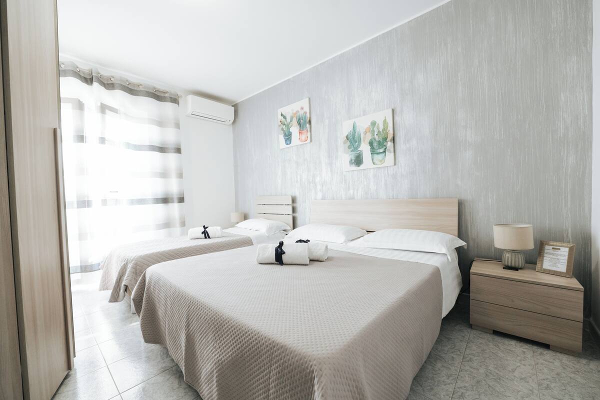 Quarto de hotel com duas camas, uma mesinha de cabeceira de madeira, uma luminária e duas obras de arte na parede.