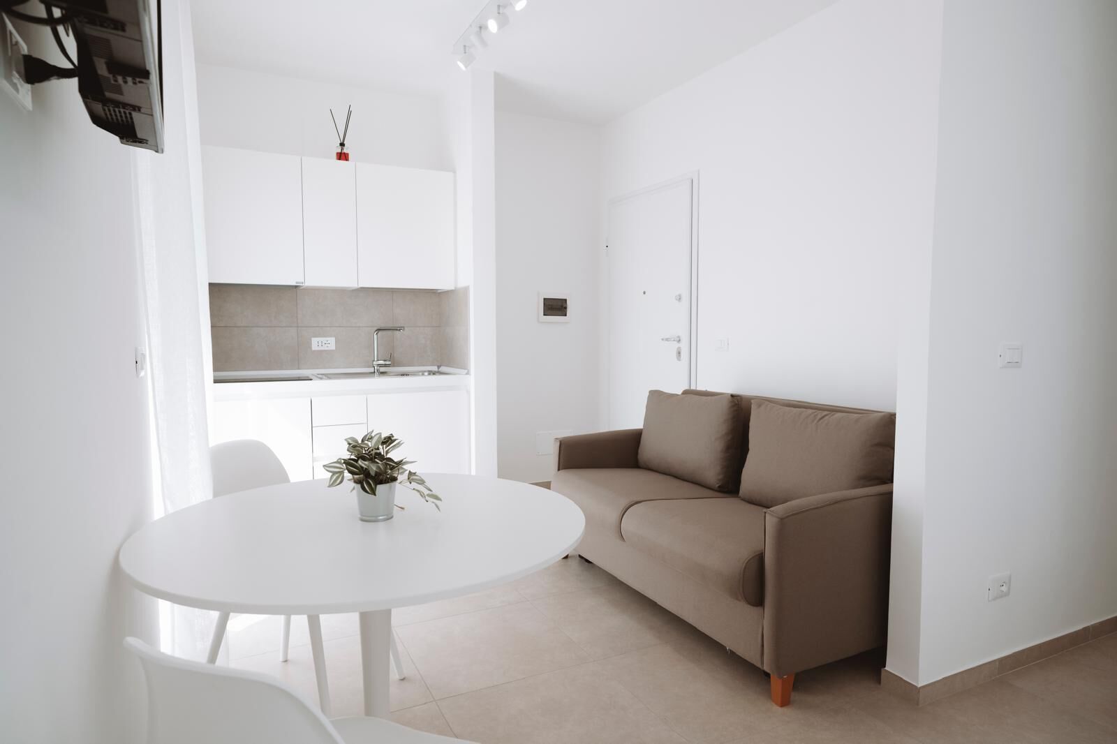 A modern, minimalist living space with a white dining table, a brown sofa, and a kitchen area with white cabinets and a tiled backsplash.