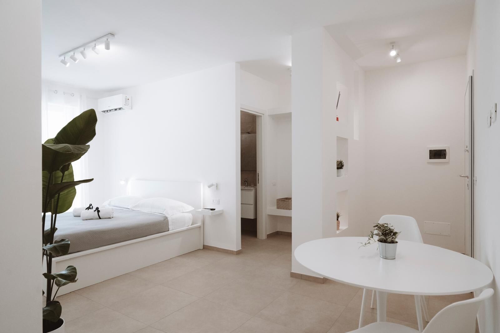 A modern, minimalist bedroom with a bed, bedside table, and a small table with a plant.