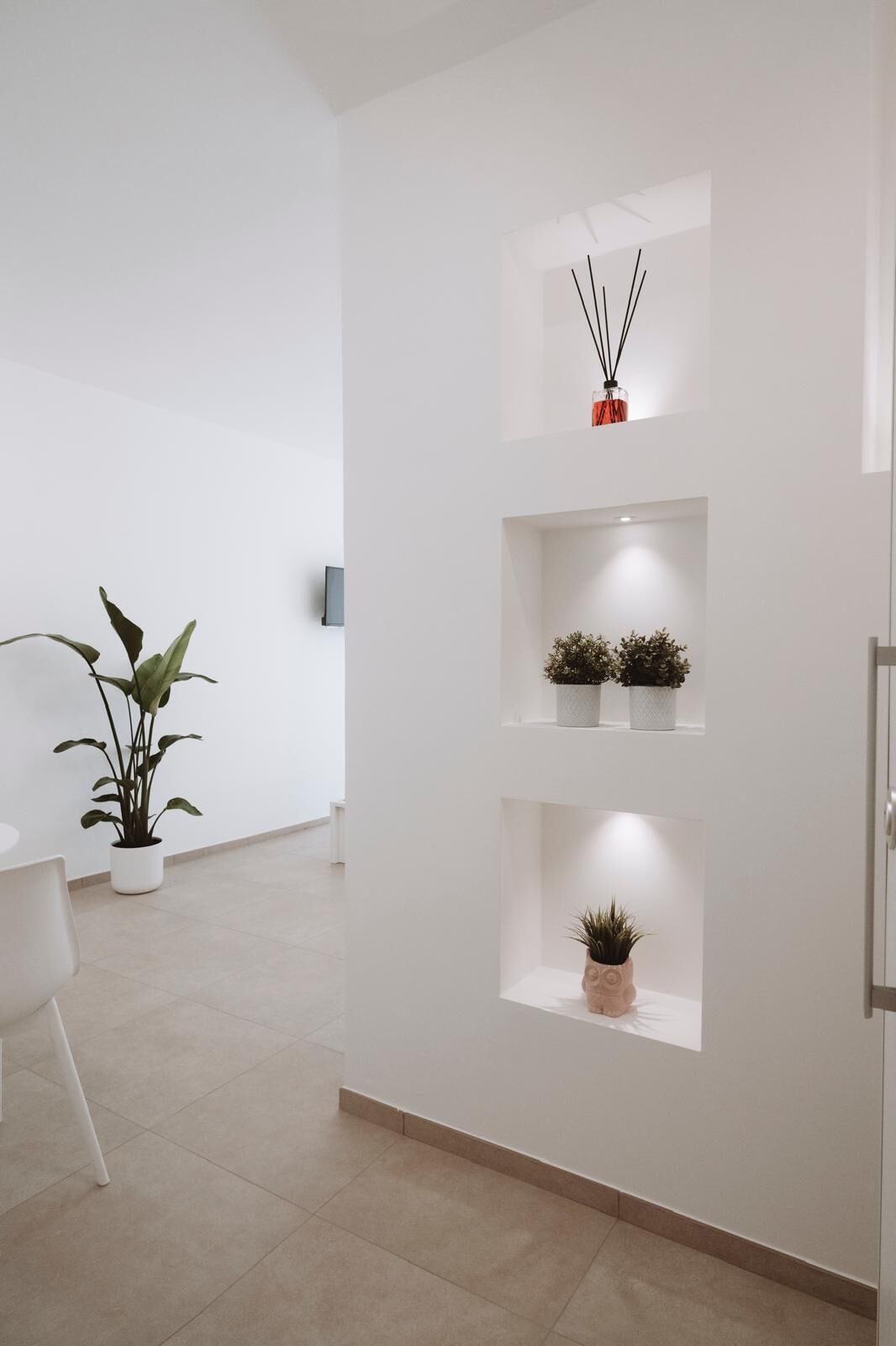 A modern, minimalist interior with white walls, a tiled floor, and three shelves with plants and a diffuser.