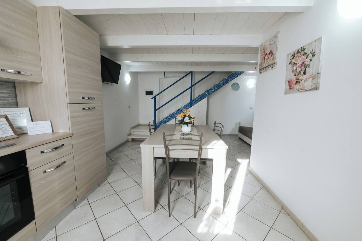 Cozinha compacta com mesa de jantar, cadeiras e escada que leva a um andar superior.