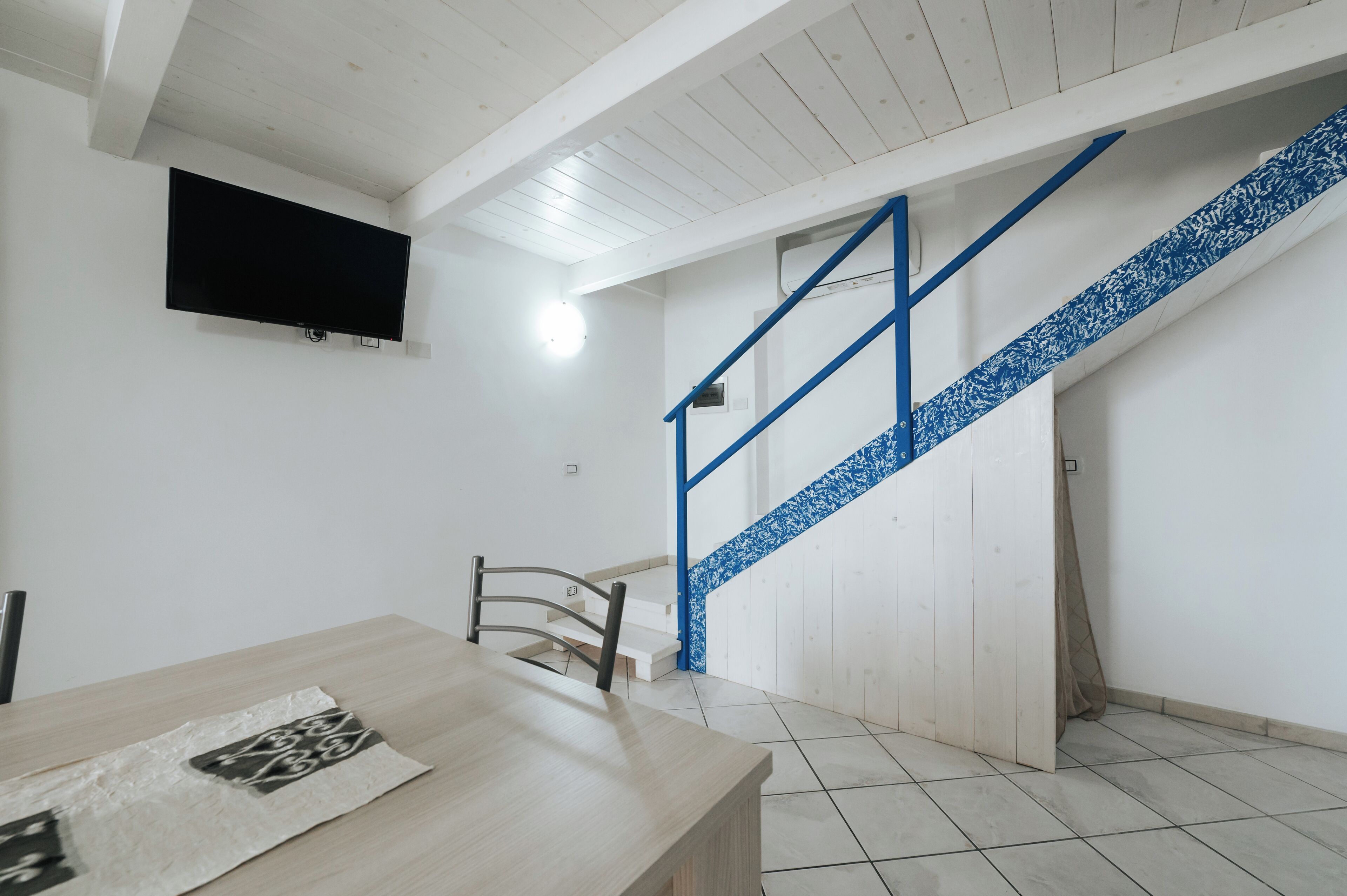 A room with a wooden table, a chair, a TV mounted on the wall, and a staircase with blue railings.