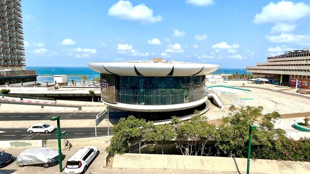 Un edificio moderno con fachada de vidrio curva, situado cerca de una playa con aparcamiento y una carretera.