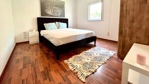 Un dormitorio con cama, mesita de noche, una obra de arte colgada en la pared, ventana y alfombra estampada.