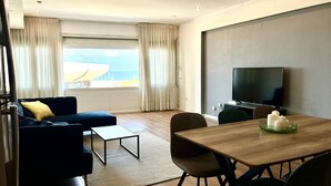 Una sala de estar moderna con un sofá, una mesa de centro, una mesa de comedor, sillas, un televisor y vistas al exterior.