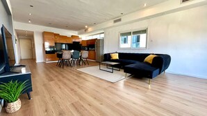Una sala de estar moderna con un sofá oscuro, una mesa de centro y un televisor. Hay una zona de comedor con una mesa y sillas. El ambiente tiene piso de madera y un ventanal grande.