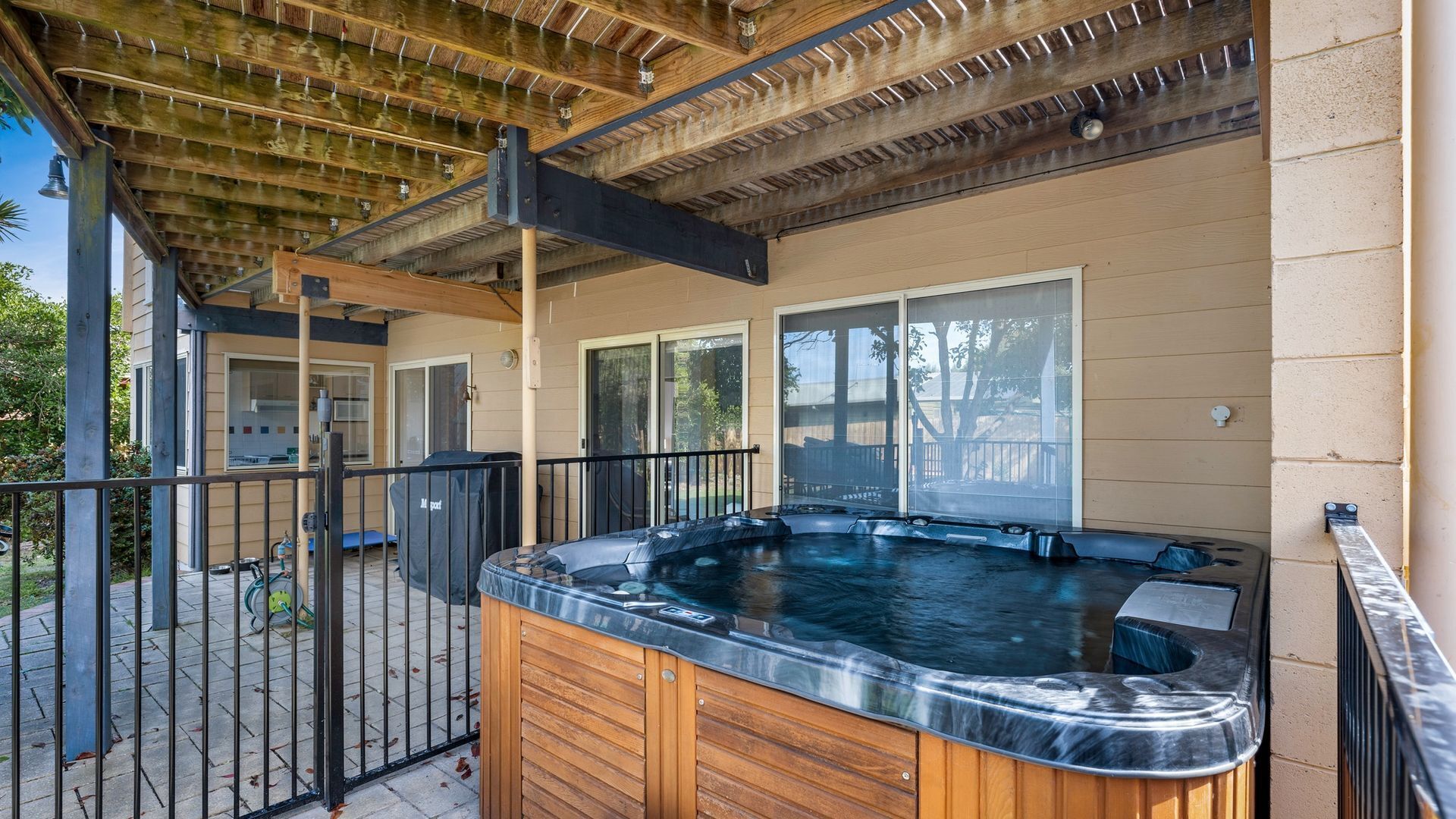 Een hottub met houten buitenkant, een pergola erboven en uitzicht op een huis met schuiframen.