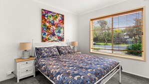 Un dormitorio con cama, dos mesitas de noche, una lámpara auxiliar y una ventana con persianas, además de un cuadro abstracto y colorido en la pared.