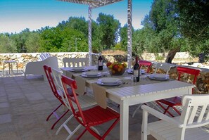 Zona pranzo all'aperto con un muro in pietra, sedie bianche e rosse e un tavolo apparecchiato per quattro persone.