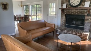 A living room with a leather sofa, a round glass coffee table, a brick fireplace, and a large wall clock. - Cute home, great for MCRD families (entire home) (Beaufort)