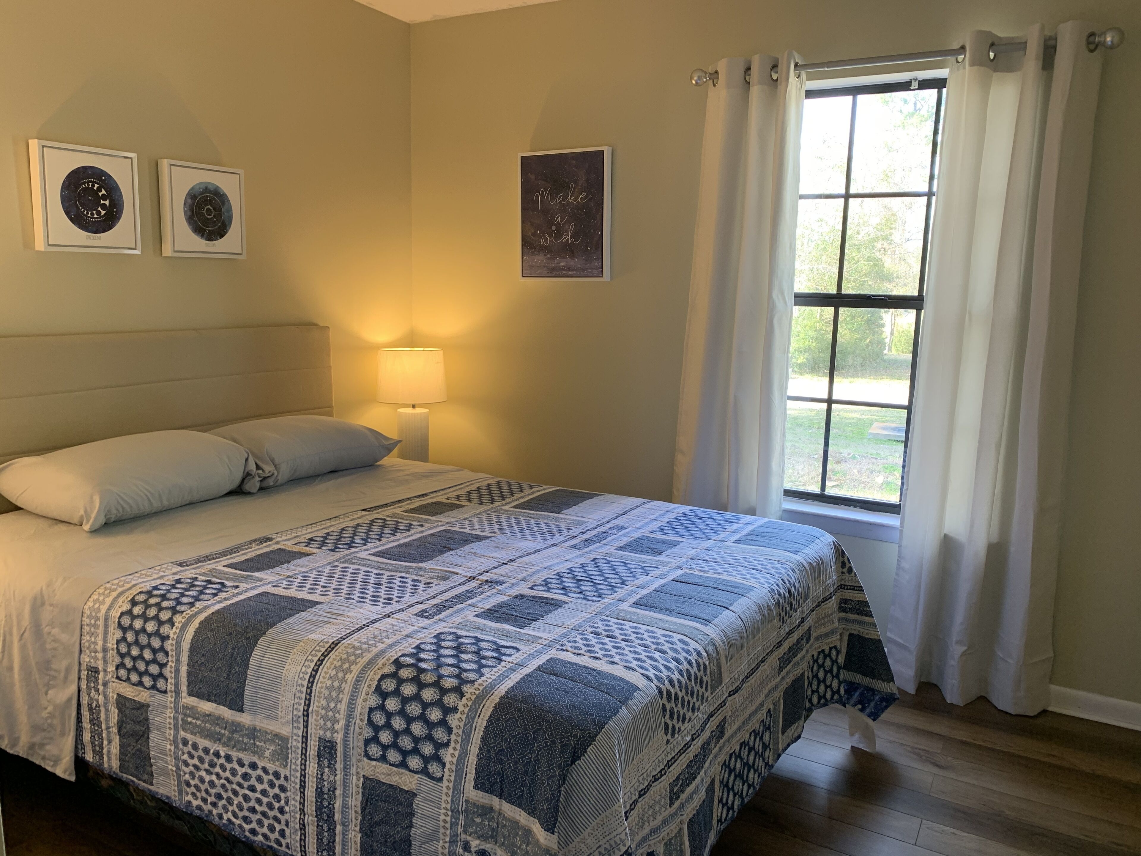 A bedroom with a large bed, a nightstand with a lamp, a window with curtains, and two framed pictures on the wall.