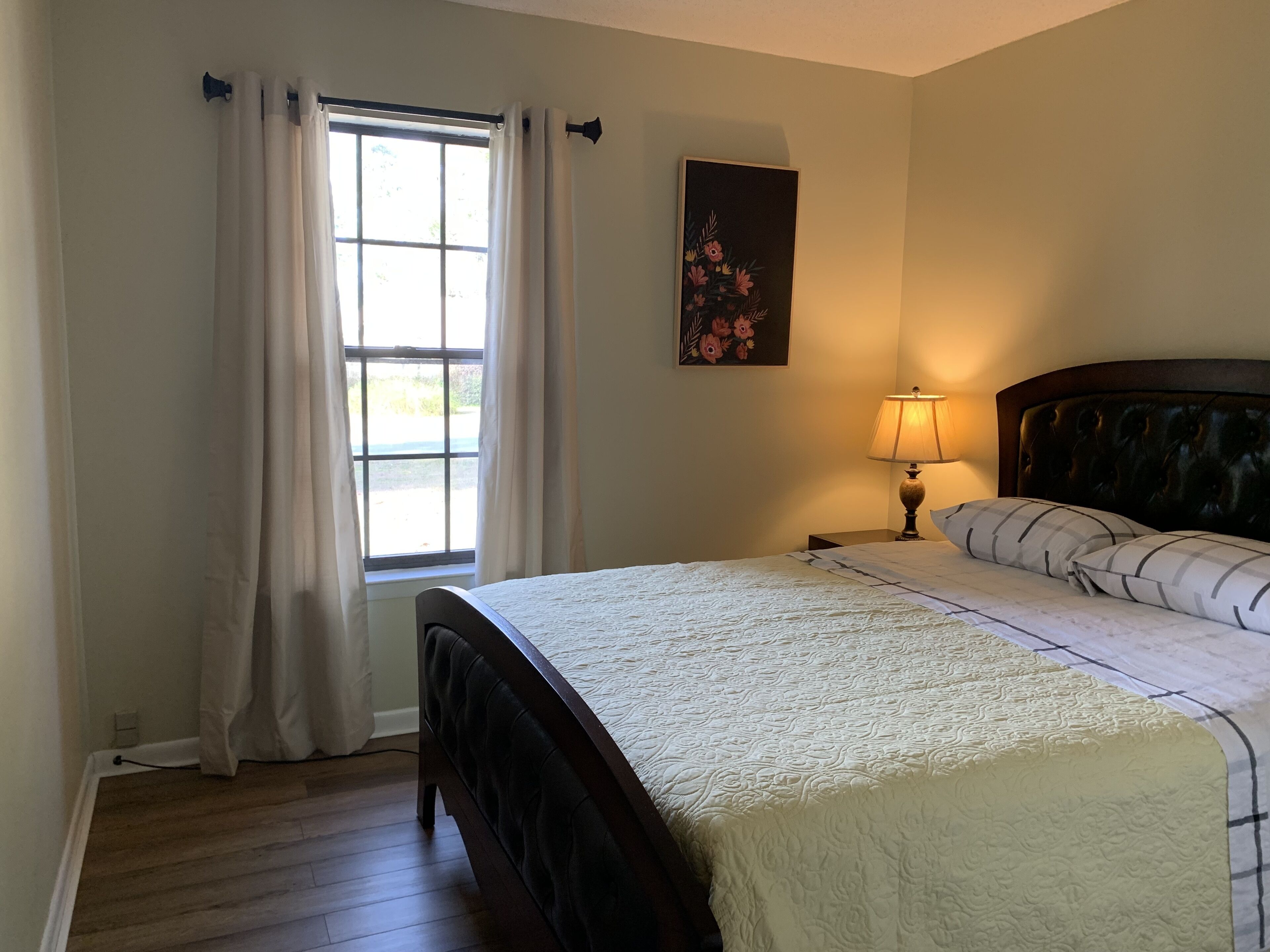 A bedroom with a bed, a window with curtains, a nightstand with a lamp, and a floral painting on the wall.