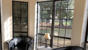 A spacious room with two black leather chairs, a small round table with a lamp, and large windows offering a view of trees and a yard. - Cute home, great for MCRD families (entire home) (Beaufort)