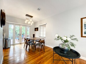 A dining area with a wooden table and chairs, a black side table with a vase of flowers, and a ceiling light fixture.