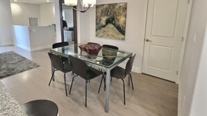 A dining area with a glass table, four chairs, a chandelier, and a painting on the wall. - Frank's Pismo Beach House: Steps to Sand & Surf (Pismo Beach)