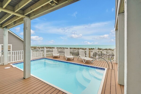 A swimming pool with a wooden deck, white lounge chairs, and a view of the ocean.