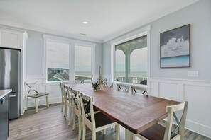 A dining area with a wooden table and chairs, large windows with a view of the ocean, and a painting on the wall. - 3 Waters Retreat: Oceanfront, 7BR 5.5BA, Pool, Elevator, EV Charger, Dog Friendly, Sleeps 20 (Oak Island)